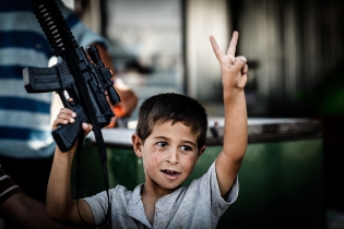  A young refugee flashes the V-sign and plays with a toy machine gun ine the Domiz camp